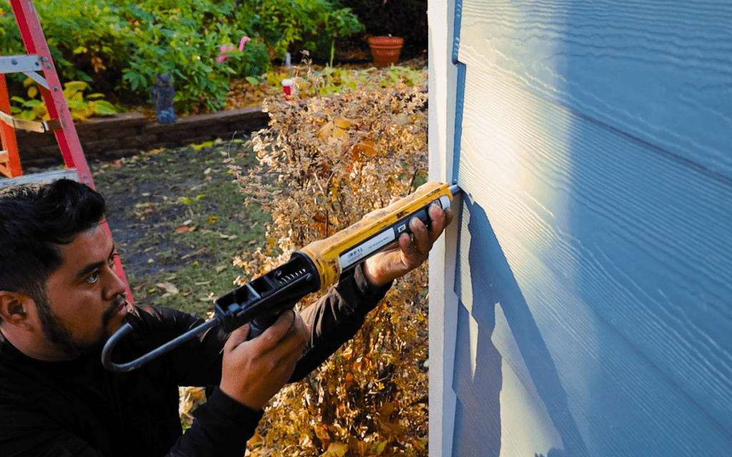 A person applies caulk with a caulking gun along the edge of blue house siding—a key step in siding damage repair—with autumn foliage and a red ladder visible in the background.