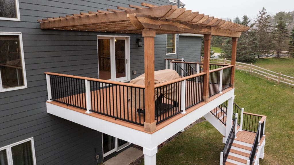 A raised wooden deck attached to a gray house features a pergola, black railings with white posts, and stairs leading down to a grassy yard. Patio furniture is covered with a beige cover. Trees and a fence are in the background.