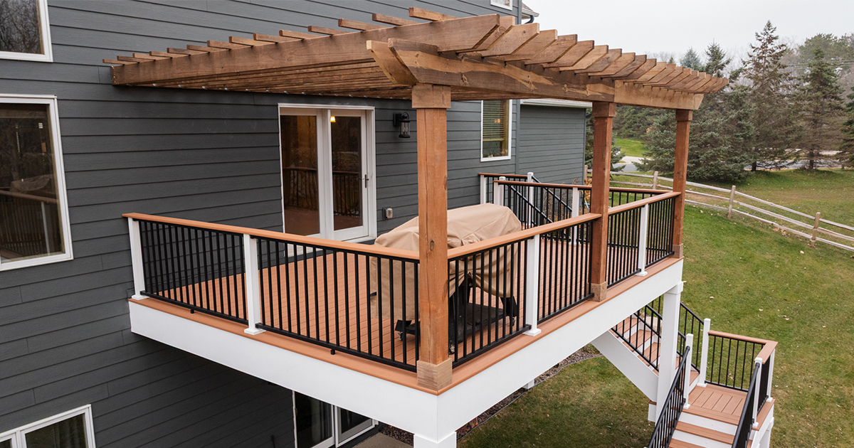 Timber Tech Cypress Deck Installation with a second-story deck with a wooden pergola, black and white railing, and covered furniture, attached to a gray house with white trim, overlooking a grassy yard with trees and a fence.