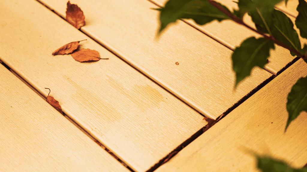 Close-up of light-colored Timber Tech Cypress decking with a few scattered brown leaves and green leafy branches partially visible on the right side, showcasing the details of this quality deck installation. Low-maintenance deck ideas from Timber Tech.