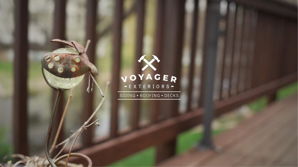 A rustic metal garden ornament stands beside a wooden deck railing, blurred in the background. The logo "Voyager Exteriors" with crossed hammers and "Siding • Roofing • Decks" highlights your trusted Twin Cities deck contractor.