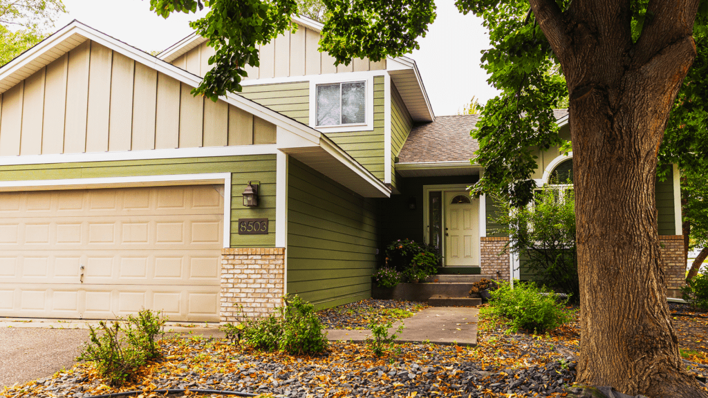 A green two-story suburban house with beige trim, an attached double garage, and a front porch features Timber Tech Cypress deck installation. A large tree shades the yard, while fallen leaves cover parts of the ground and walkway.