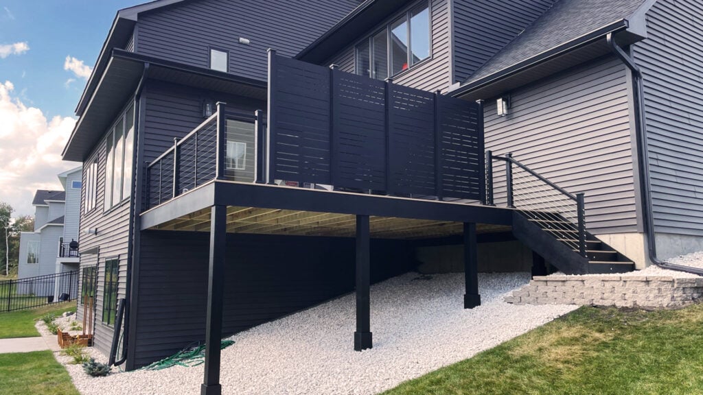 A modern dark gray house boasts a raised back deck installation with sleek metal railings, Timber Tech Cypress privacy panels, and stairs descending to a landscaped yard with white gravel and retaining walls.