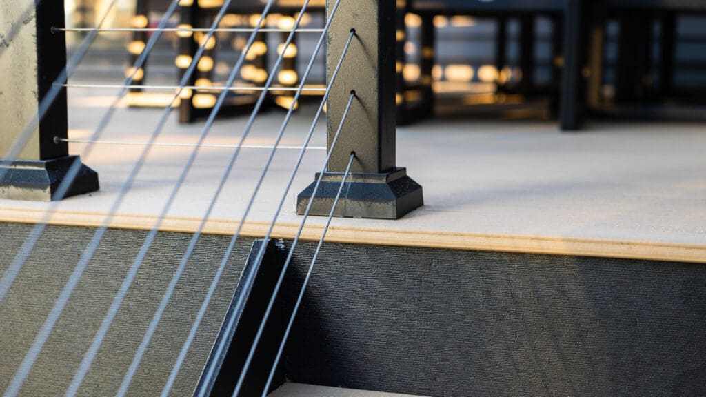 Close-up of modern outdoor deck steps featuring Timber Tech Cypress boards, metal cable railing, and sturdy posts, with sunlight highlighting the textures of the wood and metal surfaces—a showcase of expert deck installation.