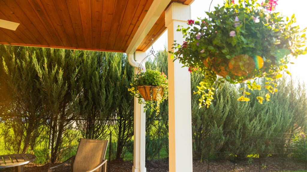 A sunlit patio with a wooden ceiling, white pillars, hanging flower baskets, a brown chair, and a round table. Greenery and tall shrubs line the background—perfect inspiration for custom deck additions or deck upgrades Twin Cities homeowners love.
