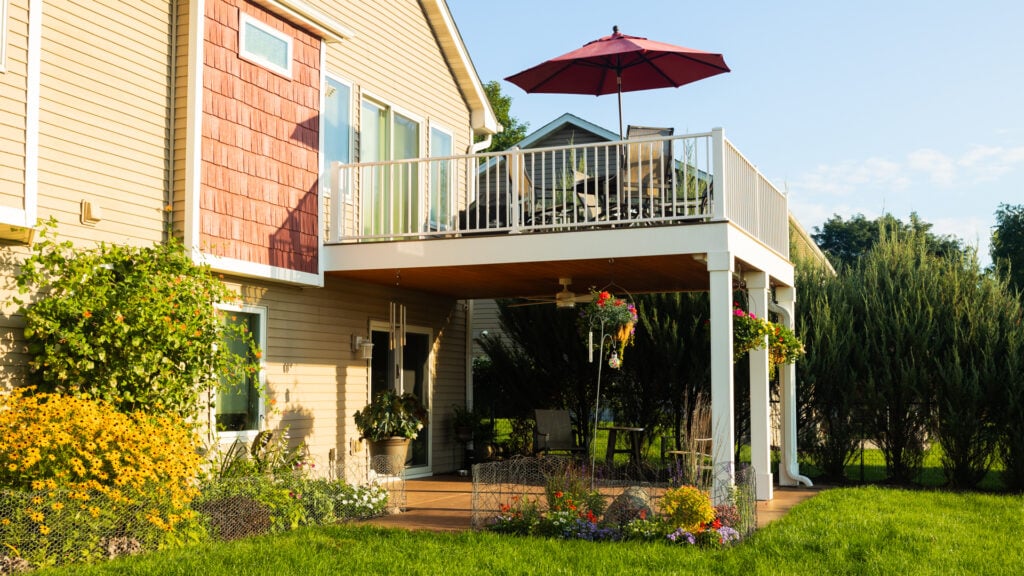 A two-story house with a spacious upper deck featuring custom deck additions like a table and red umbrella; below is a shaded patio area with plants, surrounded by a green lawn and colorful flowers on a sunny day.