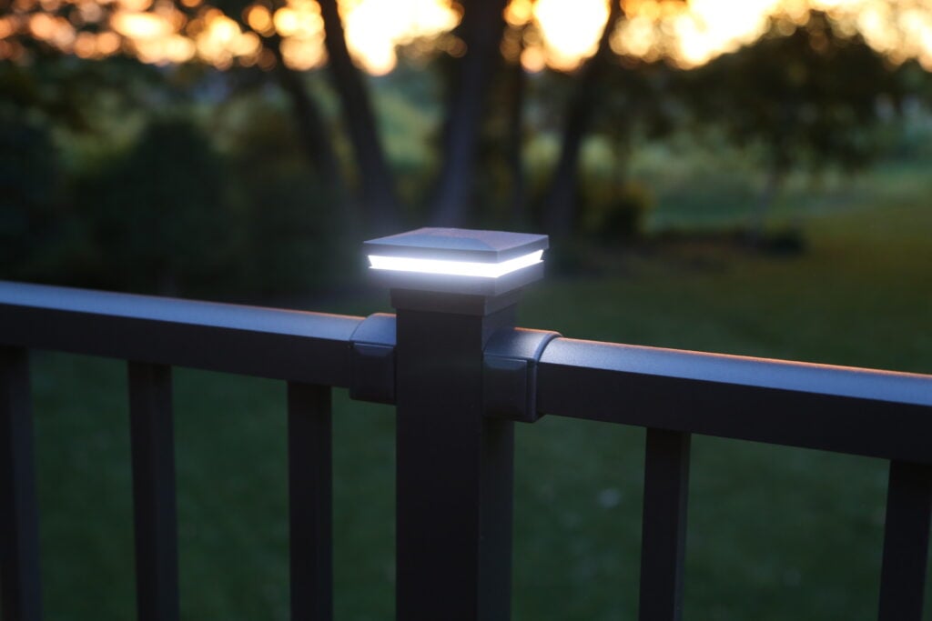 A close-up of a metal deck railing with integrated deck lighting glowing at dusk—an example of custom deck additions. Out-of-focus trees and a grassy yard are visible in the background.