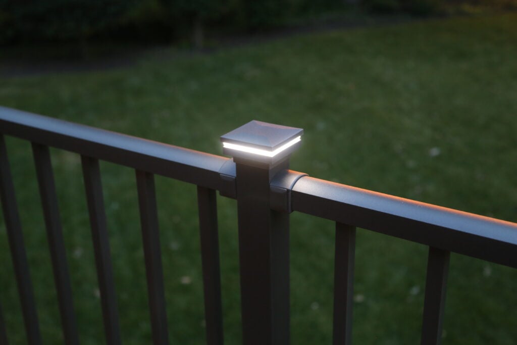 A close-up of a metal fence with a square post cap light glowing softly at dusk, custom deck additions in view, and a grassy lawn in the background.