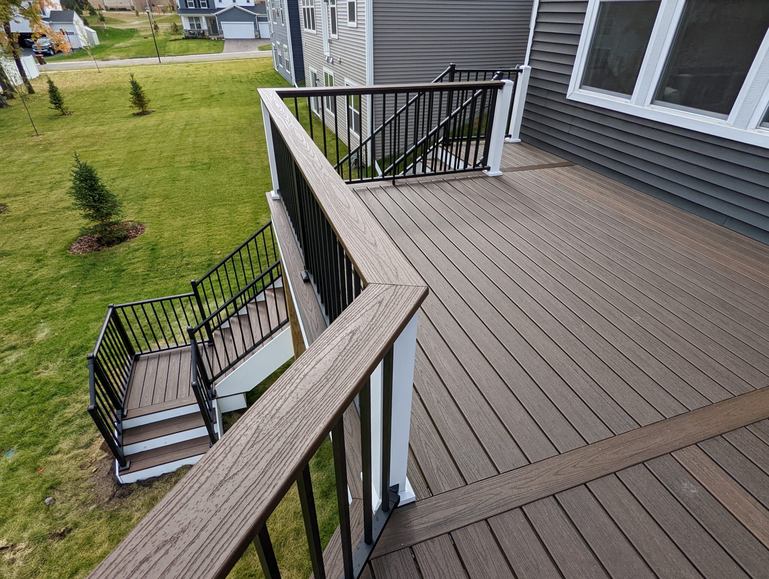 Drink rails on a modern brown composite deck with black and white railings extends from a gray house, showcasing stylish composite deck upgrades. Stairs lead down to a green lawn with young trees in a residential Twin Cities neighborhood.