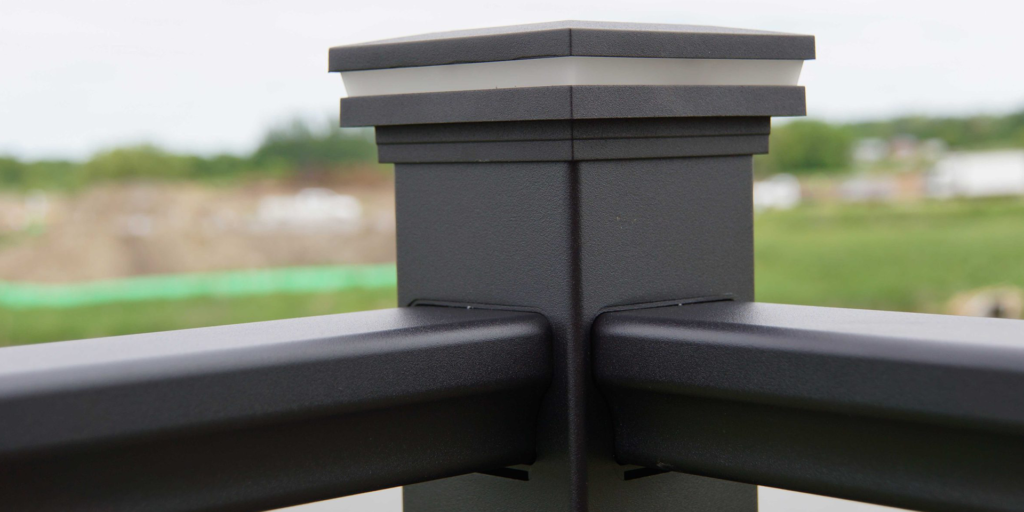 Close-up of a modern black metal fence post with horizontal rails and a square cap on top. In the blurred background with greenery, explore composite railing ideas or consider adding drink rails for extra outdoor functionality.