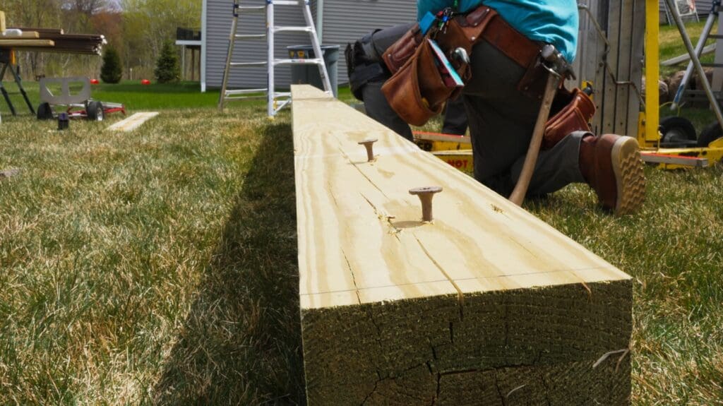 A close-up of a person kneeling on grass while securing engineered deck beams with bolts, tools in their belt and construction equipment in the background—perfect for glulam beams projects across the Twin Cities.