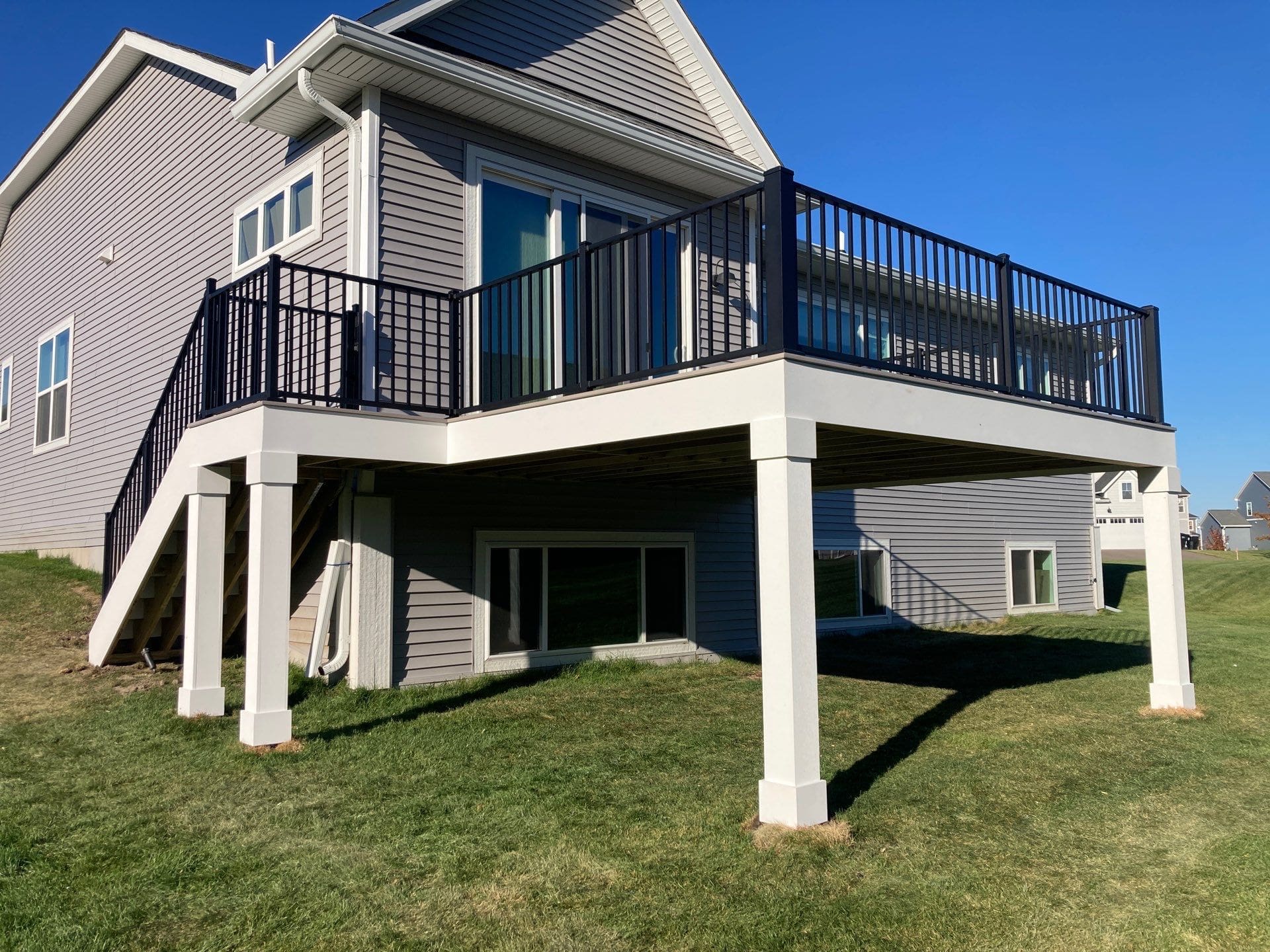 A modern house with gray siding and a raised wooden deck featuring black railings, white support columns, and engineered deck beams. Stairs lead to a grassy backyard on a sunny day in the Twin Cities.
