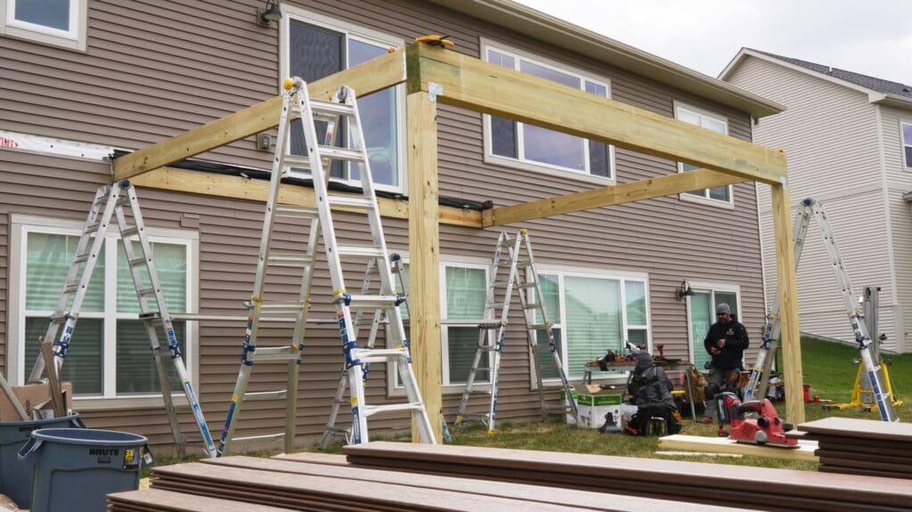 Several ladders and tools surround a wooden frame being constructed with engineered deck beams against the back of a two-story house, with two people working near the frame on a grassy yard.