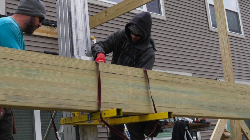 Two people work outdoors on a construction project, installing engineered deck beams. One uses a power tool while the other assists. Both are dressed in hooded jackets, and a house with siding is visible in the background.