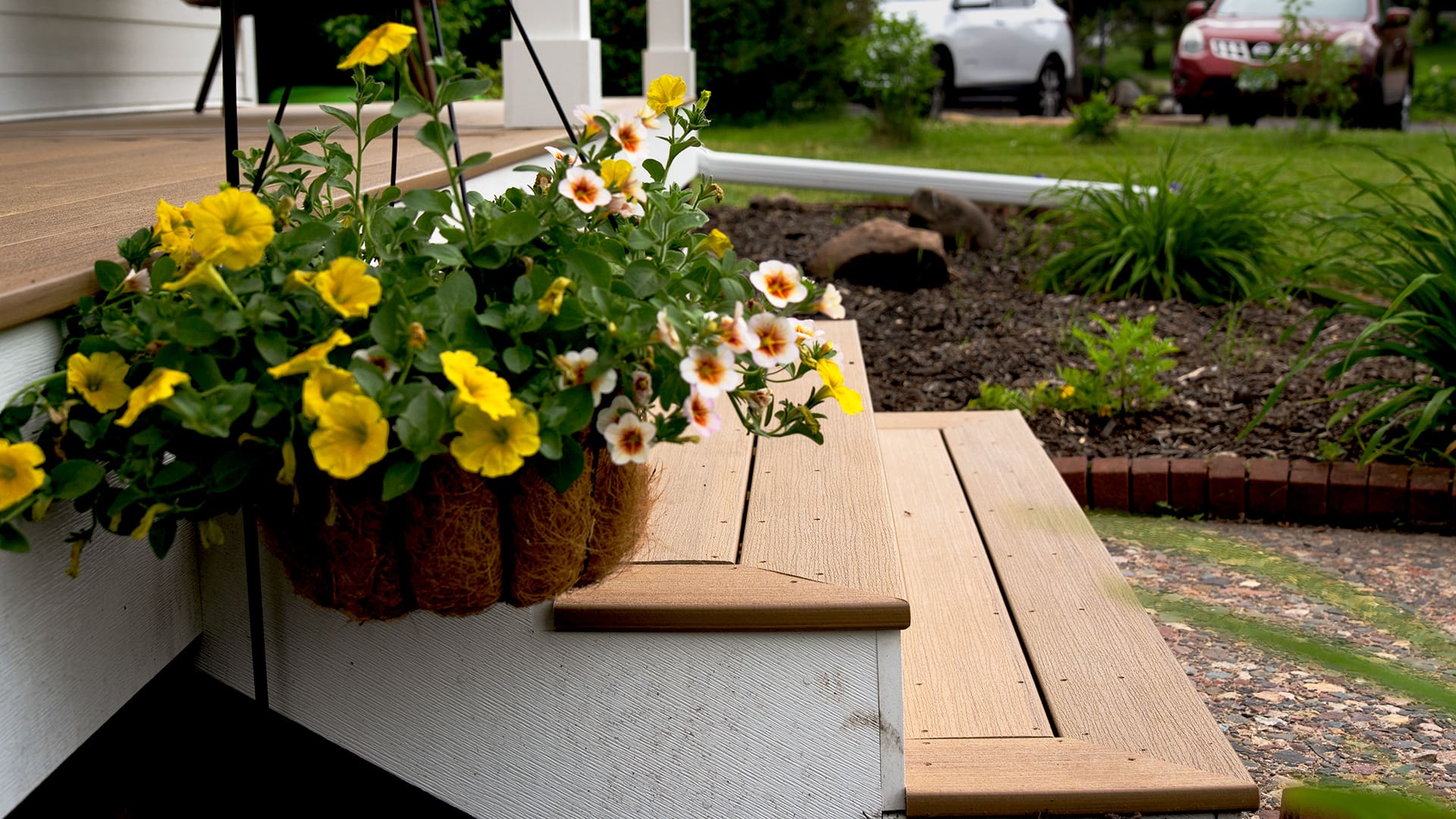 A close-up of wooden porch steps with a hanging basket of yellow and white flowers showcases a TimberTech White Oak deck. In the background, enjoy the lush garden bed, green grass, and parked cars—perfect for siding and deck installation Coon Rapids MN.
