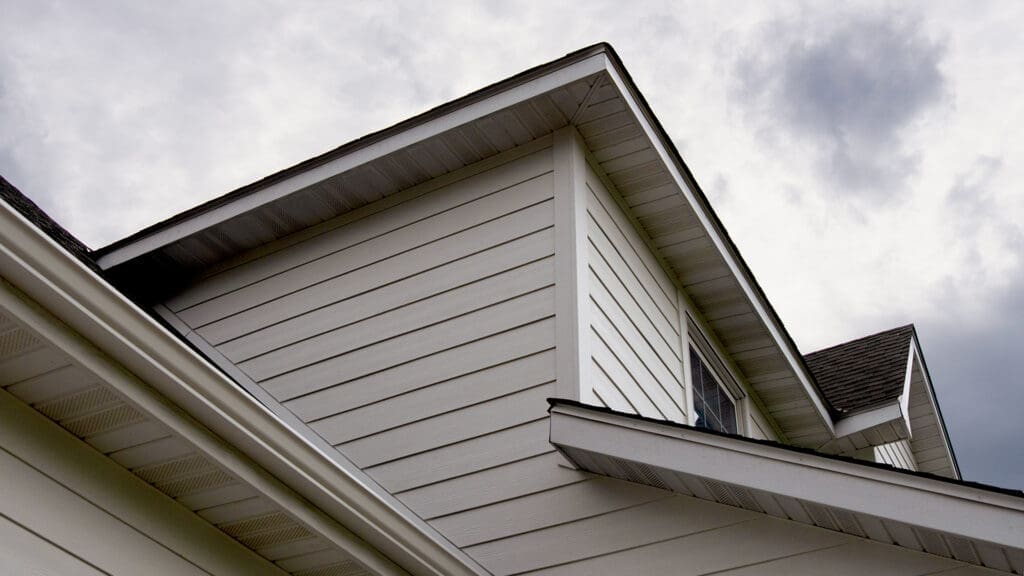 Close-up view of a house exterior featuring beige siding and sharp roof angles, with a glimpse of James Hardie Mountain Sage panels—an example of expert siding and deck installation in Coon Rapids, MN. A cloudy sky frames the scene.
