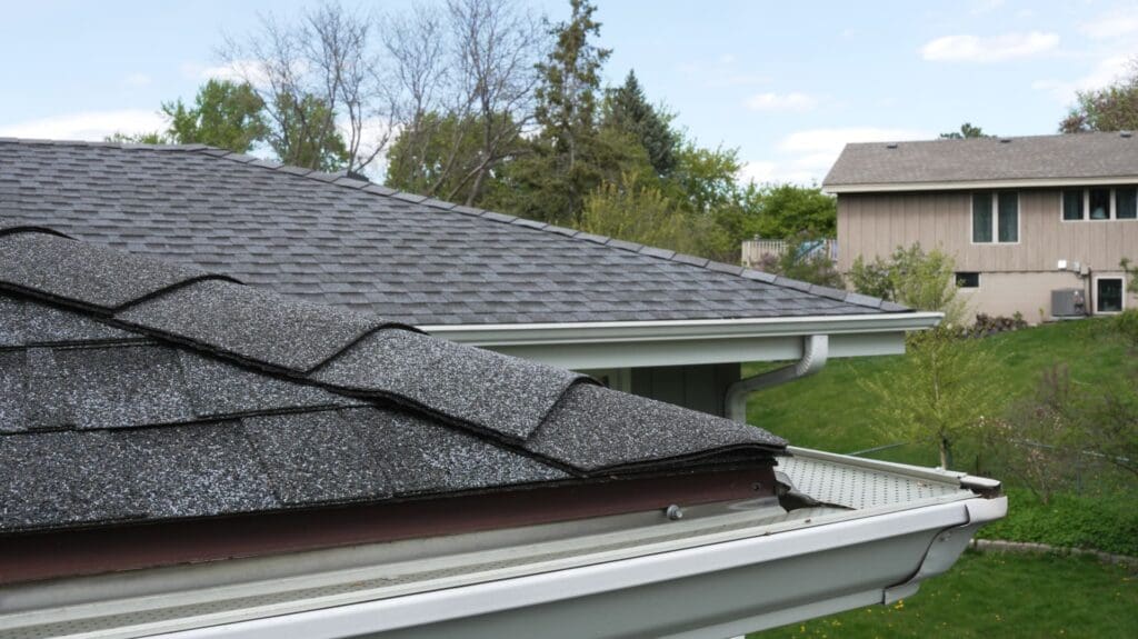 Close-up of a house roof with damaged, lifted shingles and a nearby gutter, with another house, trees, and green grass in the background—signs that could indicate exterior water leaks or water damage around windows and siding.