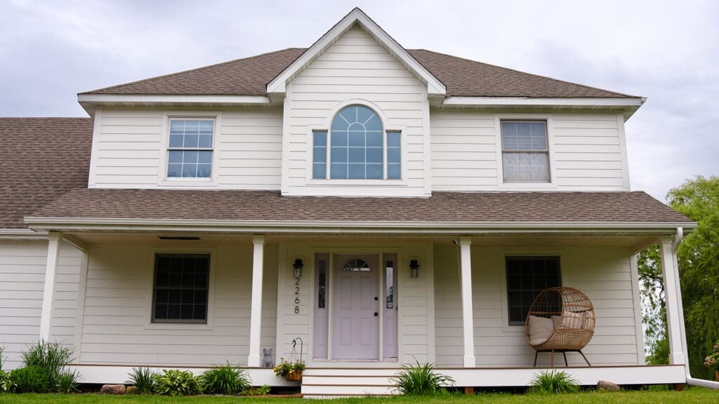 A two-story white house with a covered front porch, TimberTech White Oak deck, arched window above the entrance, gray shingle roof, and a hanging chair. Green plants decorate the front lawn—expert siding and deck installation Coon Rapids MN.