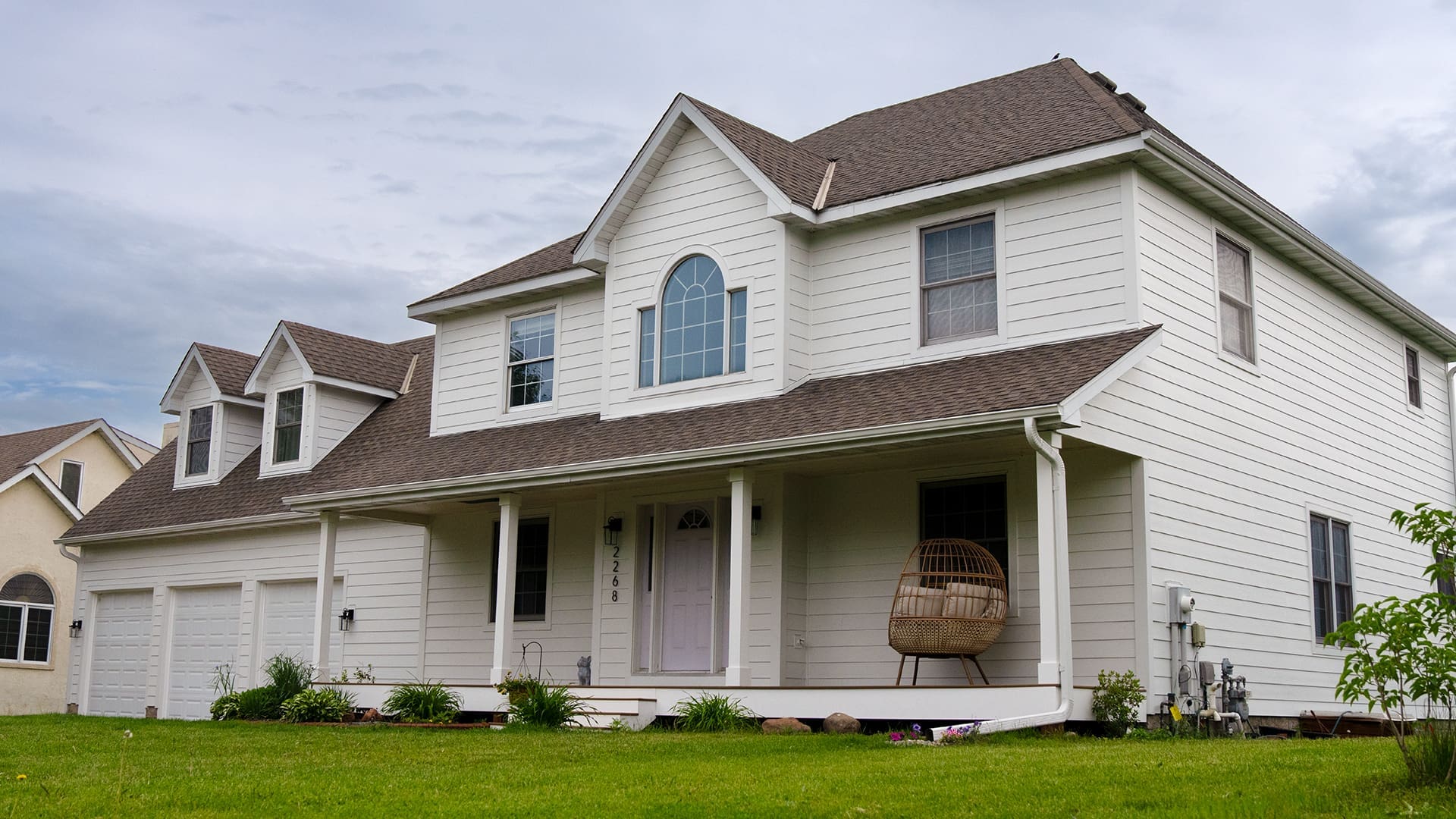 Two-story white house with a gray roof, three-car garage, and covered front porch featuring a TimberTech White Oak deck. The porch has a hanging chair, and the house is surrounded by a green lawn and small plants. Perfect for siding and deck installation in Coon Rapids MN.