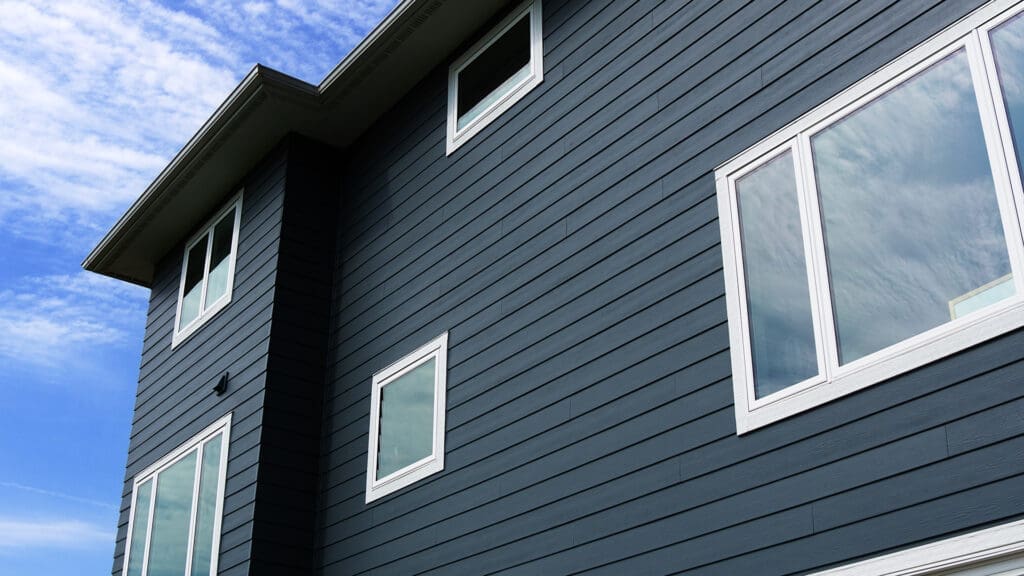 A modern house exterior with dark blue horizontal siding, large white-trimmed windows, and a cloudy sky reflected in the glass—showcasing expert siding and deck installation Coon Rapids MN. The photo is taken from a low angle.