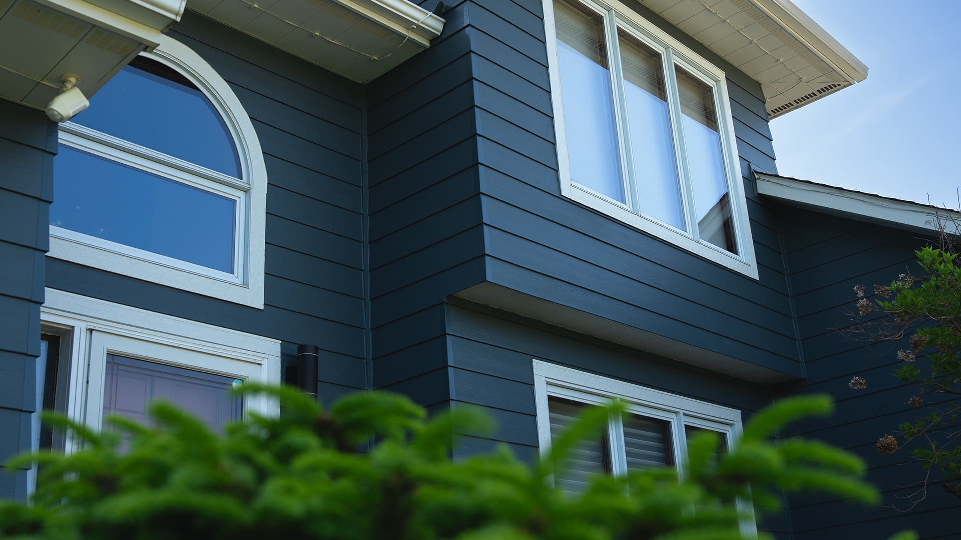 Close-up view of a modern house exterior with James Hardie Mountain Sage siding, white trim around large windows, and lush green foliage—showcasing expert siding and deck installation in Coon Rapids MN.