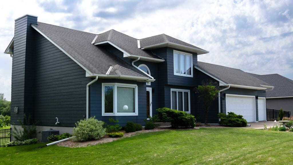 A modern two-story house with dark blue siding, white trim, and large windows features skilled siding and deck installation in Coon Rapids, MN. An attached two-car garage complements a manicured lawn under a partly cloudy sky.