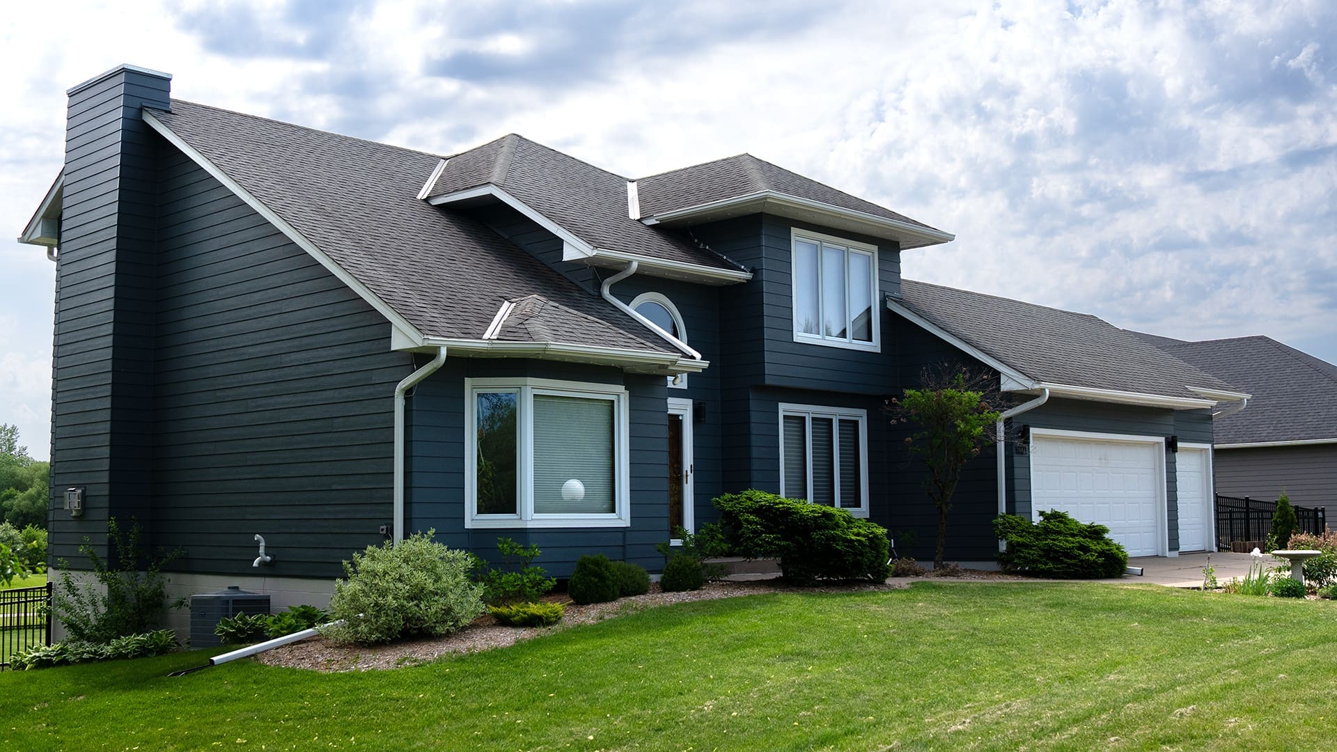 A modern two-story house with dark blue siding, white trim, and large windows features skilled siding and deck installation in Coon Rapids, MN. An attached two-car garage complements a manicured lawn under a partly cloudy sky.