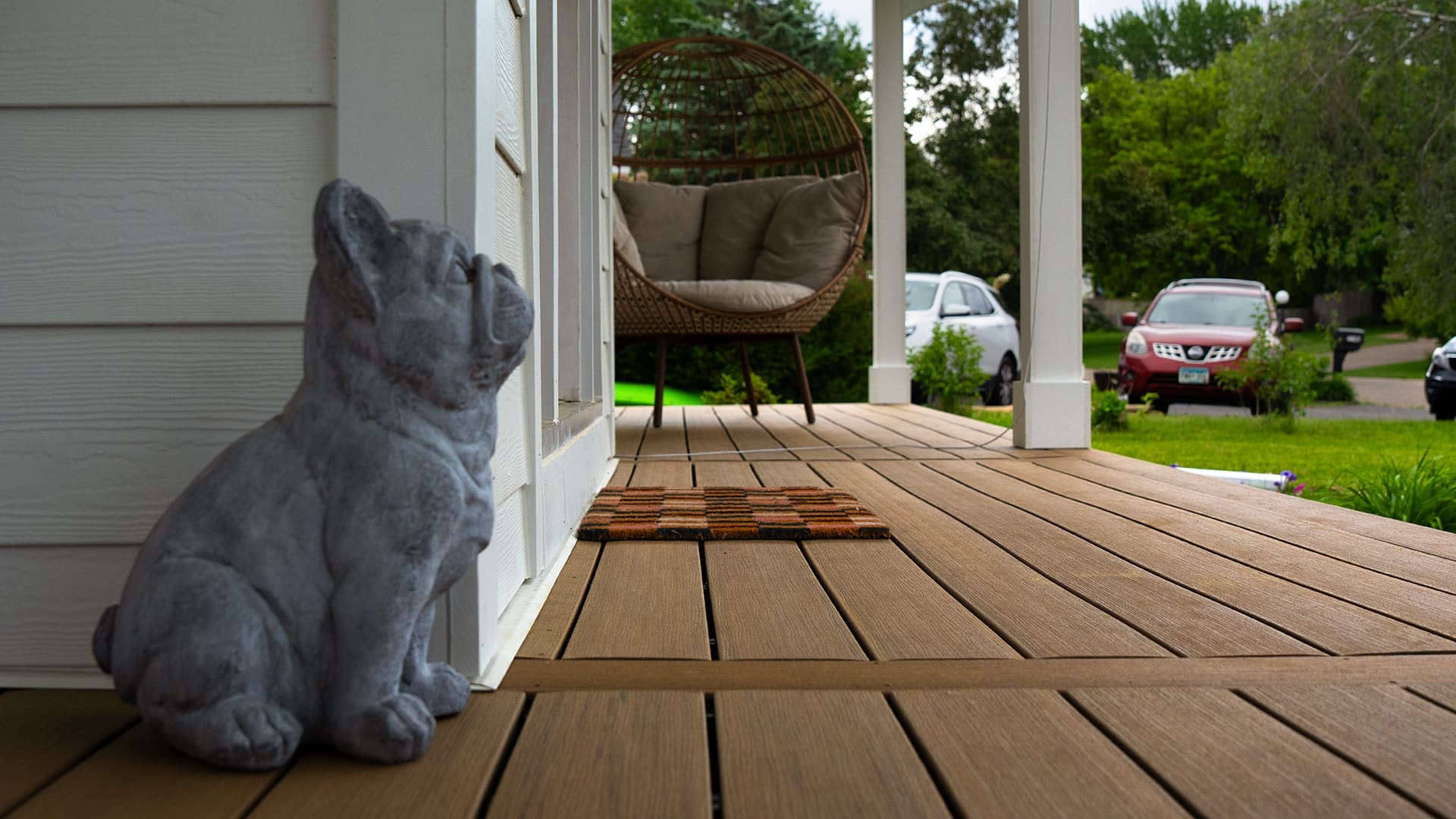 A gray dog statue sits on a wooden porch near a doormat, beside a cushioned chair. Green trees and parked cars are visible beyond the TimberTech White Oak deck in the yard.