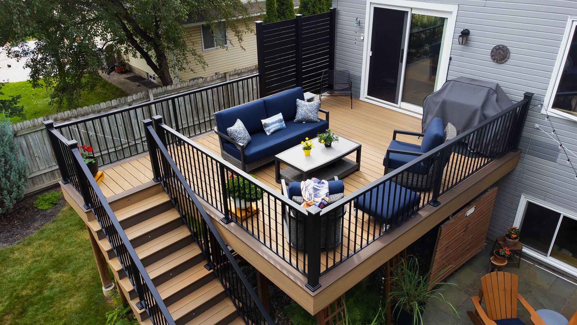 A raised deck featuring Timber Tech Cypress boards with black railings, blue outdoor furniture, a grill, potted plants, and stairs leading to a green backyard, adjacent to a gray-sided house—an inviting space showcasing expert deck installation.