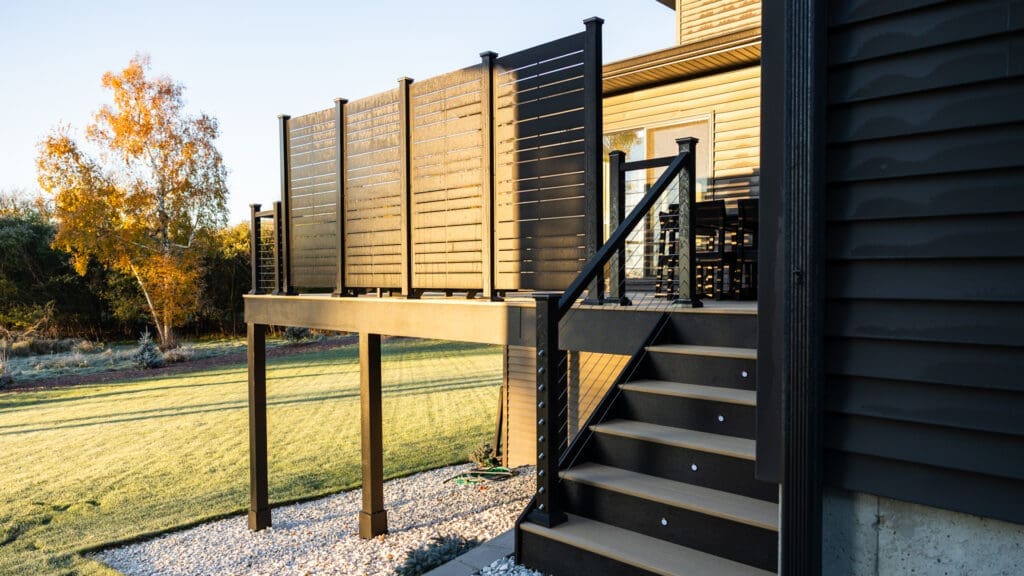 Elevated deck with privacy panels, black railing, and stairs leading to a grassy yard. Sunlight highlights the structure and an autumn tree is visible in the background—an inspiring example for those seeking low-maintenance deck ideas. Fall and winter deck construction in Minnesota — the best time of year to build a deck depends on planning and a prepared crew
