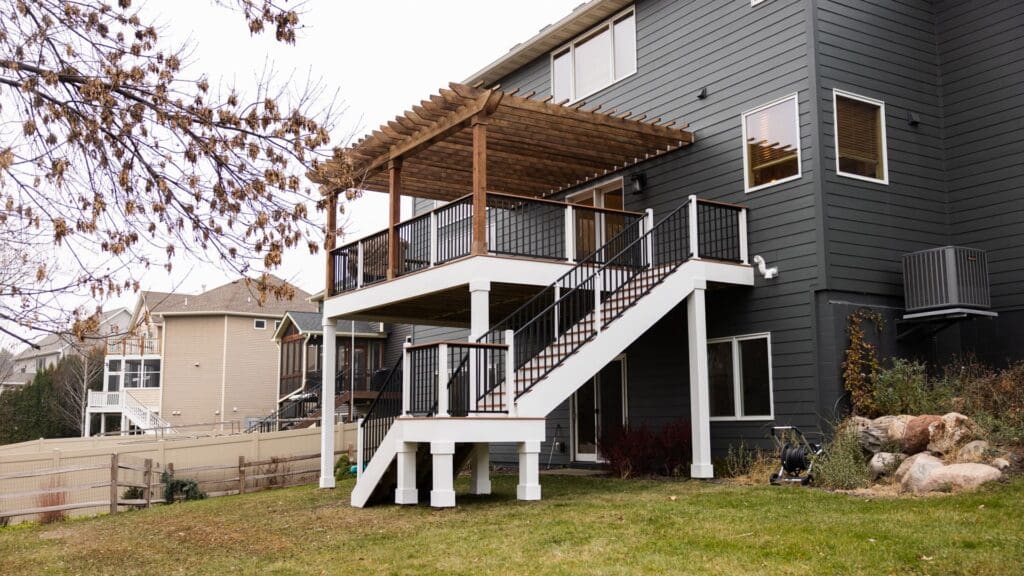 A backyard view of a modern house with a raised deck features white stairs, black railings, and a wooden pergola above the deck. The lawn is green and there are rocks and trees nearby, showcasing one of the best low-maintenance deck ideas for effortless outdoor living.
