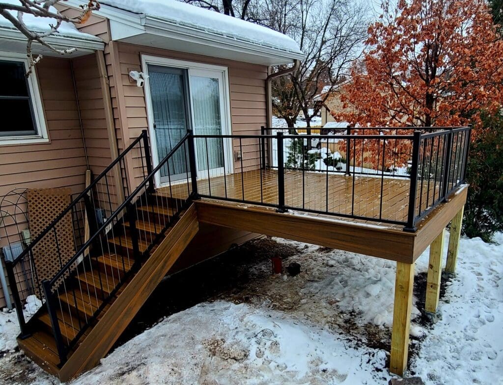 A wooden deck with black metal railings and stairs is attached to a house, covered partially with snow—a reminder that building a deck in fall can create a beautiful outdoor space ready for every season. The snowy yard below contrasts with a nearby tree adorned in orange leaves, capturing the serene atmosphere of autumn. If you’re considering such an addition, remember that obtaining a Minnesota deck permit is an important step.