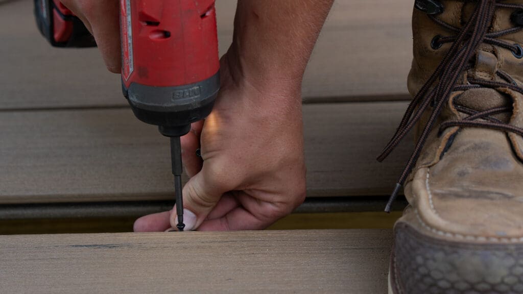 A close-up of a person’s hand using a red power drill to insert a screw into a wooden board, next to a brown work boot—a step that’s essential when preparing cold weather builds.